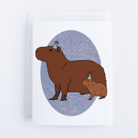 The Party Capybaras greeting card depicting an adult and baby capybara wearing pale green party hats on a pale purple oval background with confetti spots. The greeting card is laid on a white background.