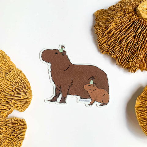 The Party Capybaras Sticker depicting an adult and baby capybara wearing pale green party hats. The sticker is laid on a white background with dried mushroom decor.